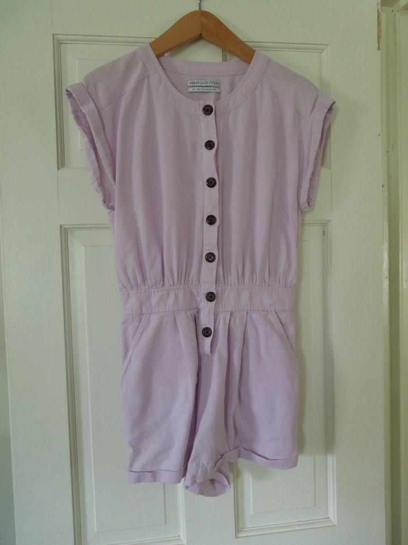 May include: A lavender button-up jumpsuit with short sleeves and a round neckline. The jumpsuit has a brand tag that reads "Urban Outfitters".