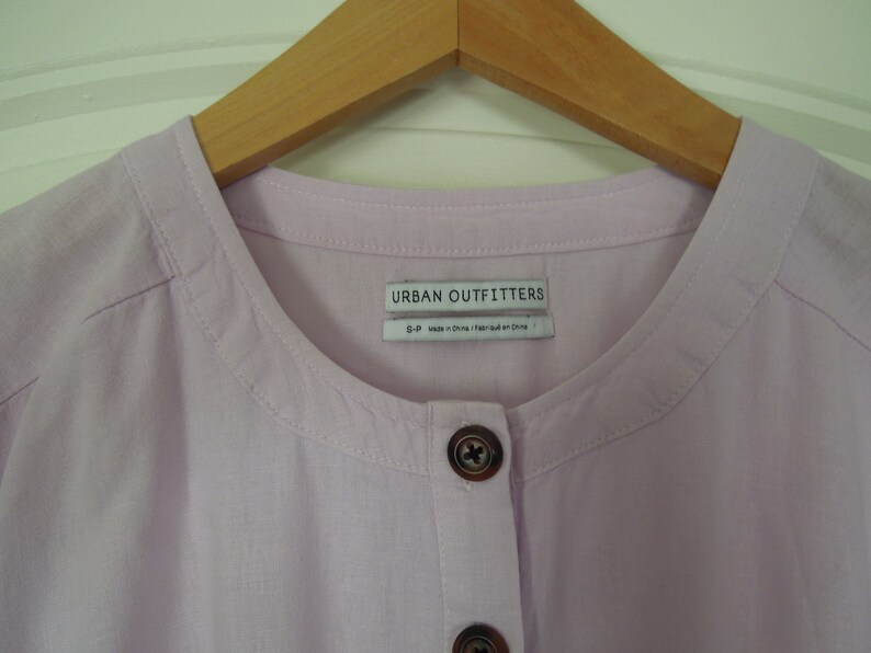 May include: A light purple button-up shirt with a round collar and a label that reads "Urban Outfitters".