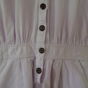 May include: A light purple button-up jumpsuit with a collared neckline and short sleeves. The jumpsuit has a gathered waist and four buttons down the front.