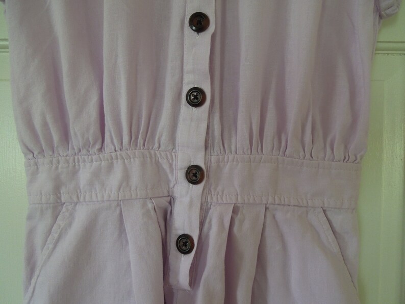 May include: A light purple button-up jumpsuit with a collared neckline and short sleeves. The jumpsuit has a gathered waist and four buttons down the front.