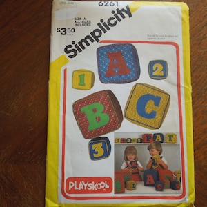 May include: Simplicity sewing pattern number 6261 for making alphabet blocks. The pattern includes instructions for making blocks with letters A, B, C, 1, 2, and 3. The pattern also includes a picture of two children playing with the blocks. The pattern is yellow with red and white text. The pattern is for sale for $350.