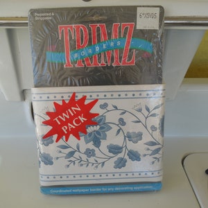 May include: A twin pack of wallpaper border featuring a floral design in blue and white. The package is labeled "TRIMZ BORDERS" and "6"X5VDS". The border is prepasted and strippable, suitable for decorating applications.