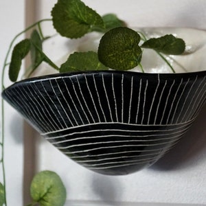 May include: A black ceramic wall planter with a white linear design. The planter is filled with green leafy plants. The planter is mounted on a white wall.