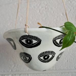 May include: White ceramic hanging planter with black eye designs. The planter is suspended by beige cords and holds a green plant. The eye pattern is repeated around the planter's exterior.