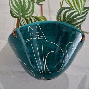 May include: A teal ceramic hanging planter with a white cat design. The planter holds a green plant with white-striped leaves. Brown leather straps suspend the planter against a white background. A charming addition to any home decor.