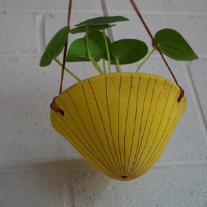 May include: A hanging planter with a bright yellow, cone-shaped pot. The pot has vertical brown lines and is suspended by brown cords. Green leafy plant sprouts from the pot. The background is a white brick wall.