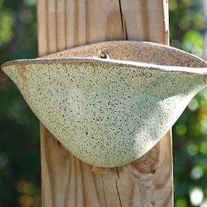 May include: A wall-mounted ceramic planter with a speckled light green and cream glaze. The vessel features a wide, curved opening and a tapered, rounded base, securely attached to a vertical wooden post with a visible grain pattern.
