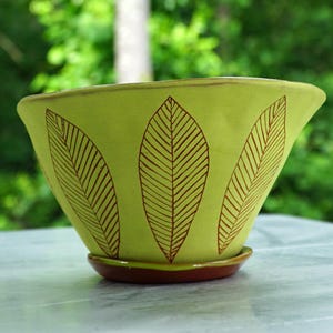 May include: A lime green ceramic planter with a flared rim and a matching saucer base. The exterior features a repeating pattern of brown etched leaf designs with detailed veins, creating a distinct contrast against the bright green surface.