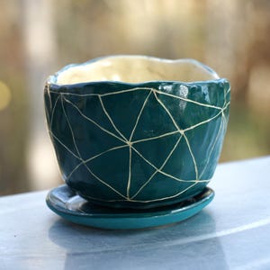 May include: A teal ceramic planter with a geometric white design, sitting on a matching saucer. The interior of the planter is a light cream color. The planter is designed for small plants and adds a decorative touch.