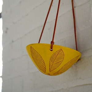 May include: A yellow hanging planter with a leaf design, suspended by brown cords against a white brick wall. The planter is bowl-shaped and features three leaf patterns. The cords are tied at the top.