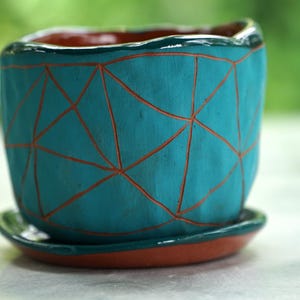 May include: A teal ceramic planter with a hand-carved geometric triangle pattern revealing the terracotta clay underneath. The pot features an uneven, organic rim and sits on a matching circular teal saucer with a terracotta base.