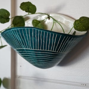 May include: A teal-colored, wall-mounted planter with white, linear detailing. The planter is filled with green leafy plants. The planter is a decorative item for indoor or outdoor use.