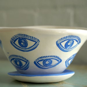 May include: White ceramic bowl with a blue eye design. The bowl has a wide, angled shape and sits on a matching saucer. The eyes are painted in a stylized, cartoonish manner, adding a whimsical touch to the piece.