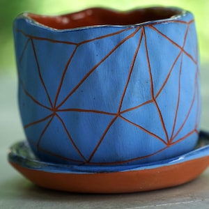 May include: A blue ceramic planter with a matte finish and a geometric pattern of recessed orange lines. The planter has a wavy, uneven rim and sits on a matching circular terracotta-colored saucer with a blue glazed edge and interior.
