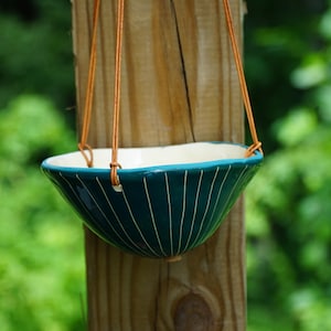 May include: A teal ceramic hanging planter with a glossy finish and vertical white pinstripes. The bowl-shaped vessel features an irregular, organic rim and is suspended by thin, light brown leather cords attached to the sides of the planter.