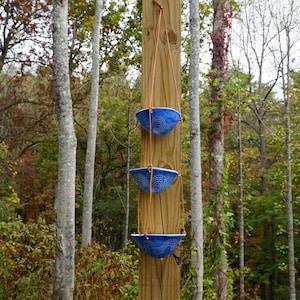 May include: Three blue ceramic hanging planters with a white swirl design are suspended from a wooden post by brown rope. The planters are set against a backdrop of trees and foliage, creating a natural aesthetic.