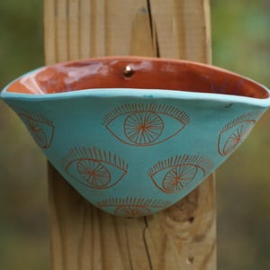 May include: A turquoise ceramic wall planter with a terracotta interior. The planter is decorated with repeating orange eye designs. It is mounted on a wooden post, showcasing a unique, handcrafted aesthetic. The planter is ideal for small plants.