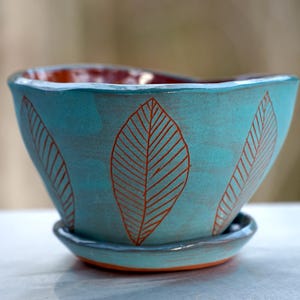 May include: A turquoise ceramic planter with a matching saucer. The planter features a leaf design in a reddish-brown color. The interior of the planter is a reddish-brown color. The planter is approximately 5 inches in diameter.