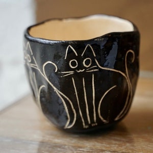 May include: A small, handmade black ceramic bowl with a glossy finish. The bowl features a cream-colored interior and is decorated with etched cat designs. The cat illustrations are in a simple, line-art style, adding a whimsical touch to the piece.