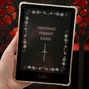 May include: A Kindle e-reader with a clear protective case. The screen displays the words "emotional support kindle" in a gothic font, framed by a floral border. A sword illustration is centered below the text. The Kindle logo is at the bottom.