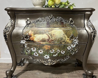 Könnte beinhalten: Eine antike Kommode aus Silber mit verzierten Details und floralen Akzenten. Die Kommode hat drei Schubladen mit einer gemalten Szene einer Frau, die in einem Blumenfeld liegt. Ein weißer Krug mit Blumendekor steht auf der Kommode.