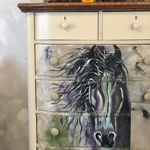 Puede incluir: Una c&oacute;moda blanca con seis cajones, cada uno con una perilla redonda. Los dos cajones superiores son lisos, mientras que los cuatro cajones inferiores est&aacute;n decorados con una imagen pintada de la cabeza de un caballo negro. La c&oacute;moda tiene una tapa de madera oscura.