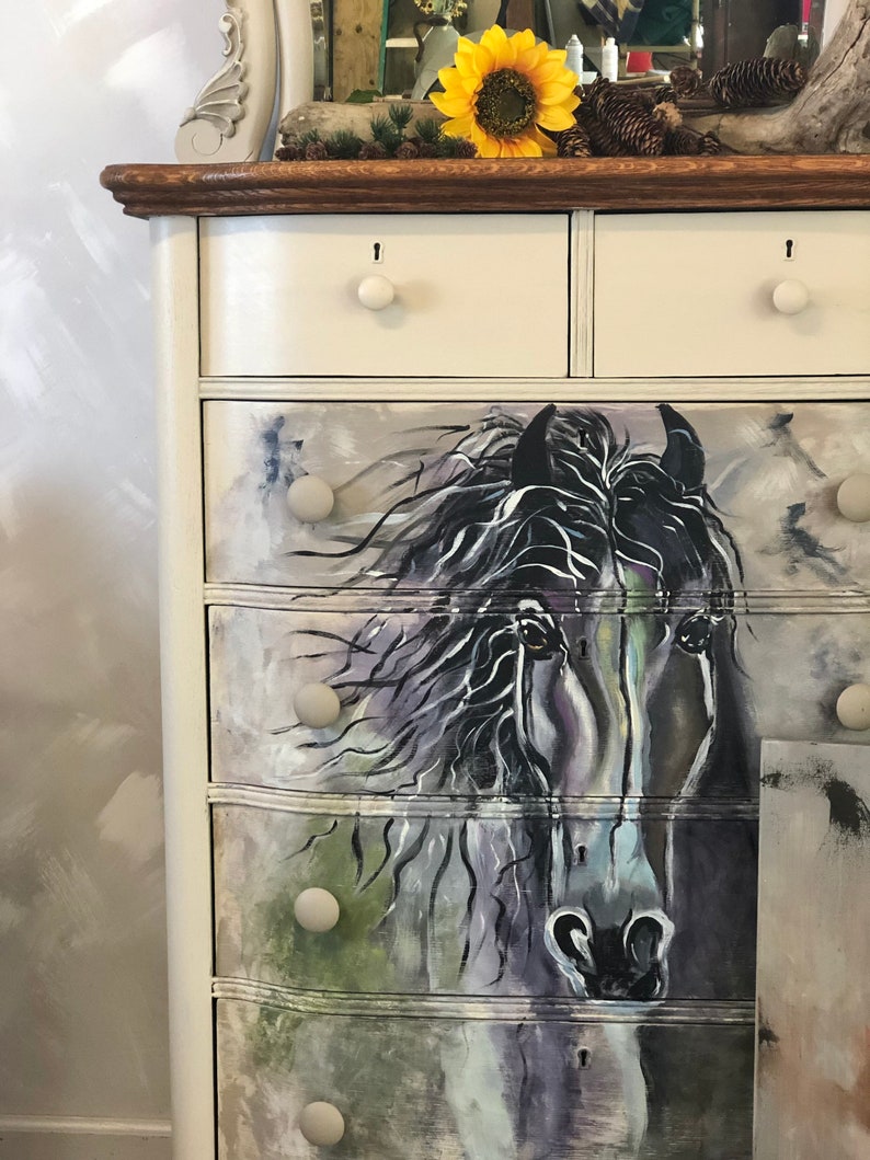 Puede incluir: Una c&oacute;moda blanca con seis cajones, cada uno con una perilla redonda. Los dos cajones superiores son lisos, mientras que los cuatro cajones inferiores est&aacute;n decorados con una imagen pintada de la cabeza de un caballo negro. La c&oacute;moda tiene una tapa de madera oscura.