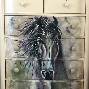 Puede incluir: Una c&oacute;moda blanca con cinco cajones que presenta una imagen pintada de la cabeza de un caballo negro. Los cajones tienen pomos redondos blancos.