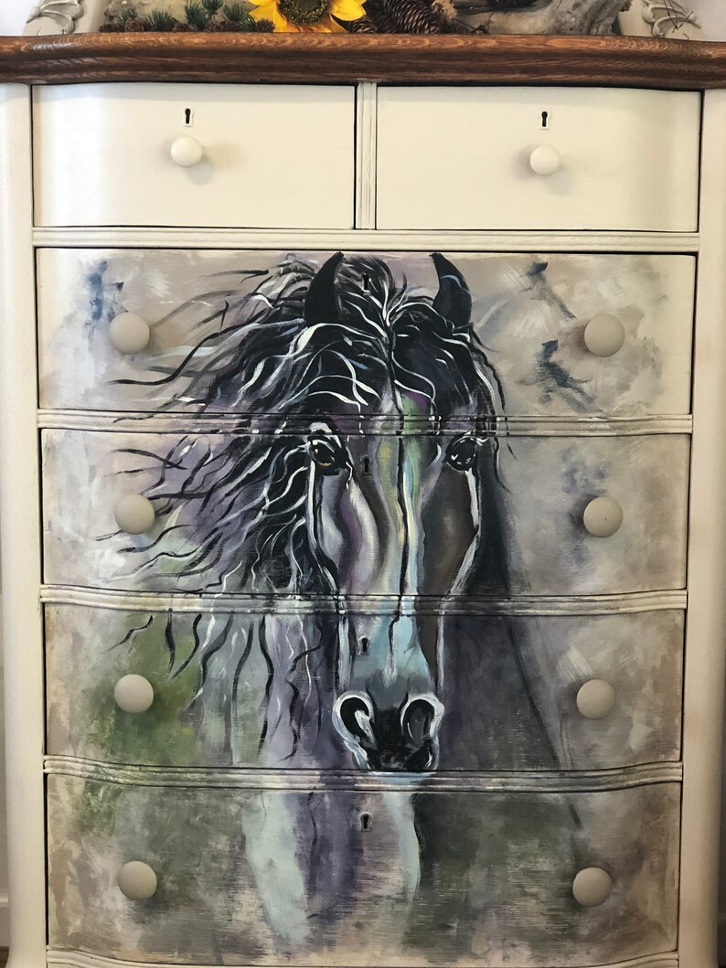 Puede incluir: Una c&oacute;moda blanca con cinco cajones que presenta una imagen pintada de la cabeza de un caballo negro. Los cajones tienen pomos redondos blancos.