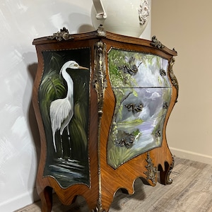Pode incluir: Um ba&uacute; de gavetas de madeira com uma cena pintada &agrave; m&atilde;o de uma gar&ccedil;a branca de p&eacute; num p&acirc;ntano do lado esquerdo e uma cena de lago do lado direito. O ba&uacute; tem detalhes em lat&atilde;o ornamentados e um vaso de cer&acirc;mica branco no topo.