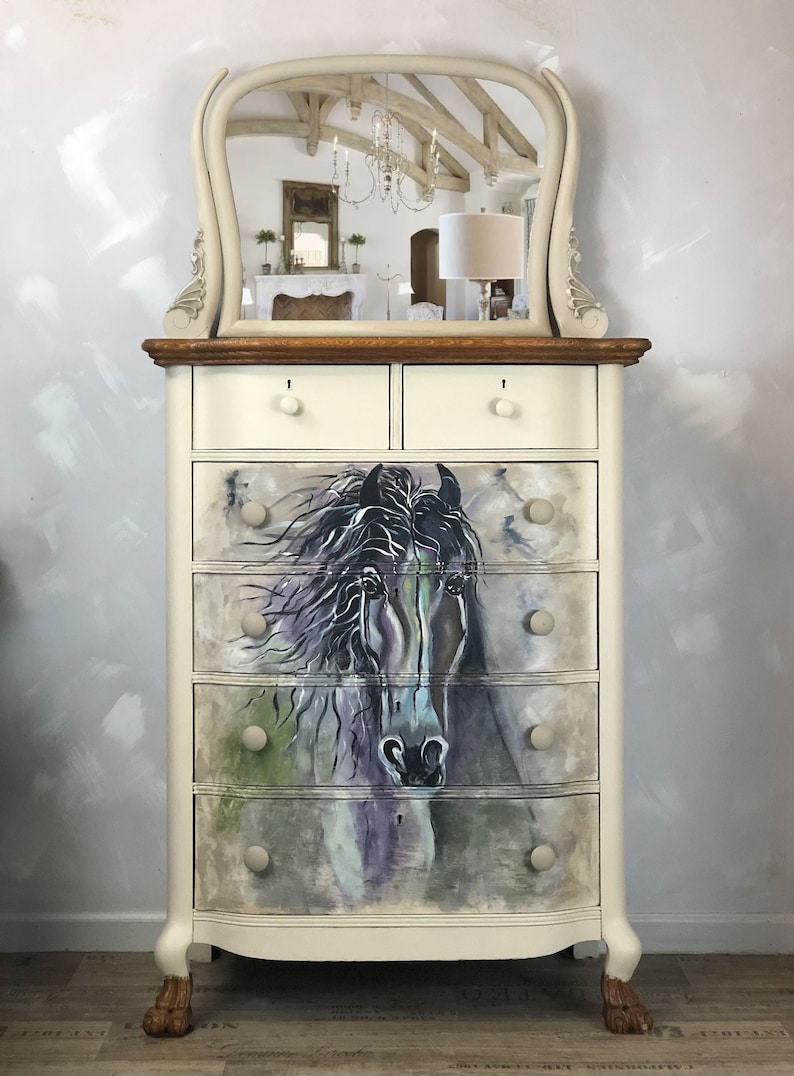 Puede incluir: Una c&oacute;moda blanca pintada con una tapa de espejo biselada. La c&oacute;moda tiene seis cajones con dise&ntilde;os de cabeza de caballo pintados. La c&oacute;moda tiene patas de madera.