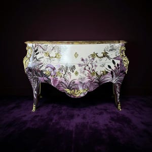 May include: A white and gold dresser with a botanical design in shades of purple and green. The dresser has ornate gold trim and legs, and a curved front. The floral pattern includes leaves, flowers, and butterflies.