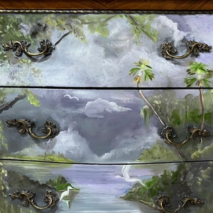 Pode incluir: Uma c&oacute;moda de madeira com tr&ecirc;s gavetas pintadas com uma cena de paisagem. As gavetas apresentam um lago, &aacute;rvores e p&aacute;ssaros. Cada gaveta tem uma pega de lat&atilde;o decorativa.