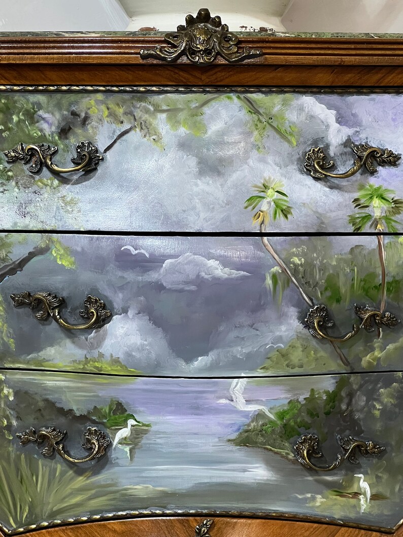 Pode incluir: Uma c&oacute;moda de madeira com tr&ecirc;s gavetas pintadas com uma cena de paisagem. As gavetas apresentam um lago, &aacute;rvores e p&aacute;ssaros. Cada gaveta tem uma pega de lat&atilde;o decorativa.