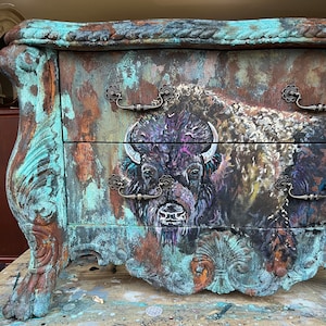 May include: A distressed teal blue dresser with two drawers features a painted image of a bison with a purple and brown color scheme. The dresser has ornate metal handles and a rustic, vintage look.
