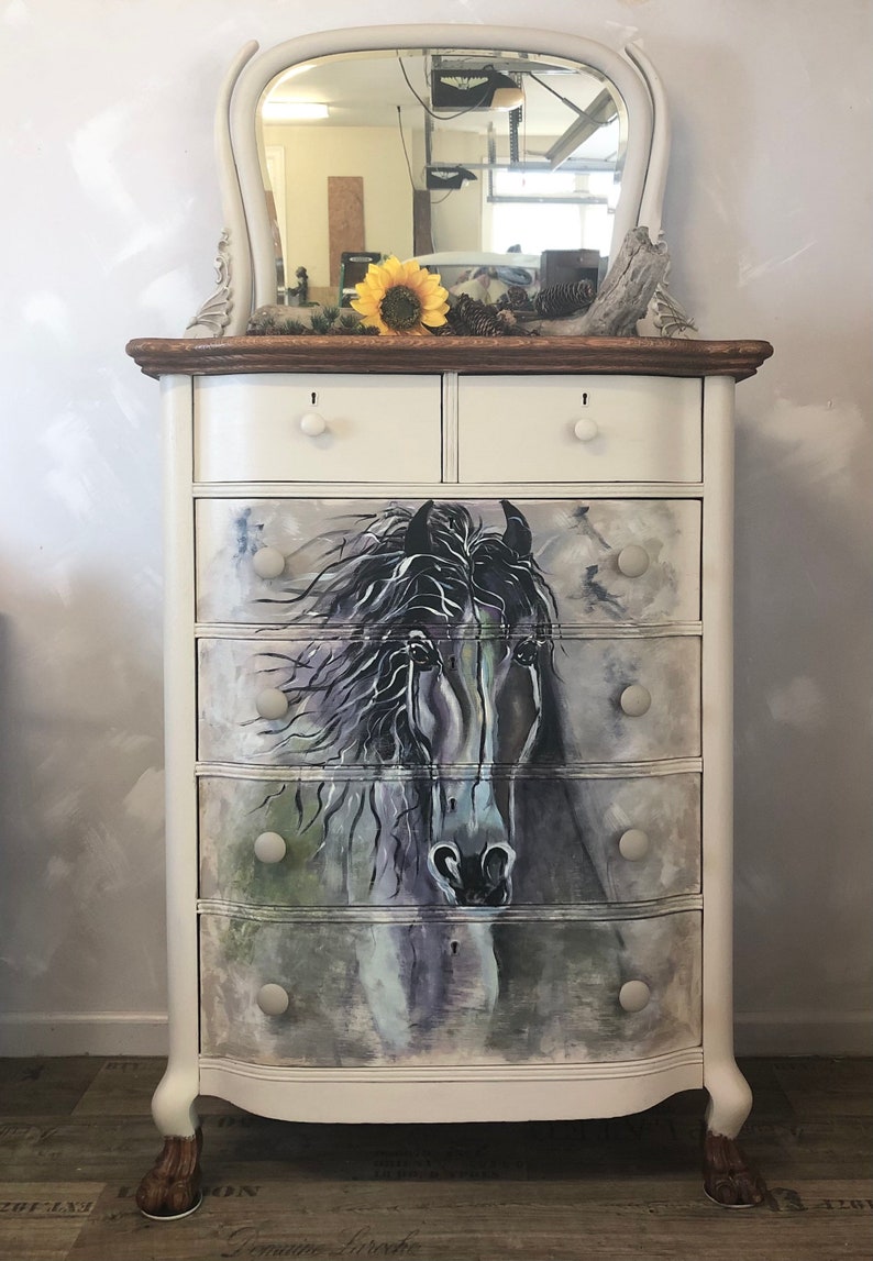 Puede incluir: C&oacute;moda blanca pintada con un espejo biselado en la parte superior. La c&oacute;moda tiene seis cajones con pomos blancos y una imagen pintada de una cabeza de caballo en la parte delantera.