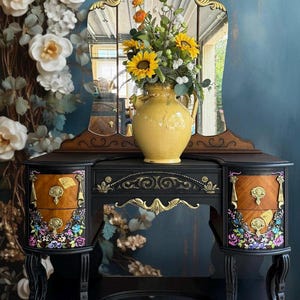 May include: An antique vanity table with a tri-fold mirror. The black table has two drawers with floral designs and gold hardware. A yellow vase with sunflowers and other flowers sits on top. The background features floral wallpaper.