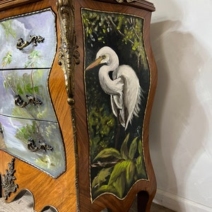 Pode incluir: Um ba&uacute; de gavetas de madeira com uma cena pintada &agrave; m&atilde;o de uma gar&ccedil;a branca numa floresta verde exuberante. O ba&uacute; tem ferragens de lat&atilde;o ornamentadas e um design esculpido na frente.