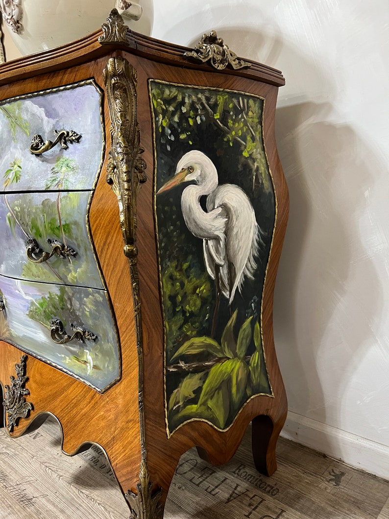 Pode incluir: Um ba&uacute; de gavetas de madeira com uma cena pintada &agrave; m&atilde;o de uma gar&ccedil;a branca numa floresta verde exuberante. O ba&uacute; tem ferragens de lat&atilde;o ornamentadas e um design esculpido na frente.