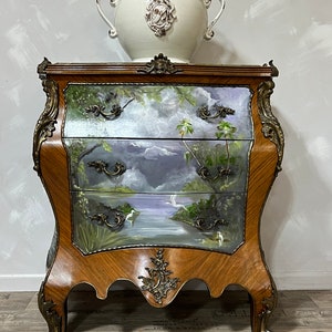 Pode incluir: Um ba&uacute; de gavetas de madeira com uma cena pintada de um lago e palmeiras. O ba&uacute; tem detalhes dourados ornamentados e est&aacute; sobre um piso de madeira. Um vaso de cer&acirc;mica branco com um design decorativo est&aacute; no topo do ba&uacute;.