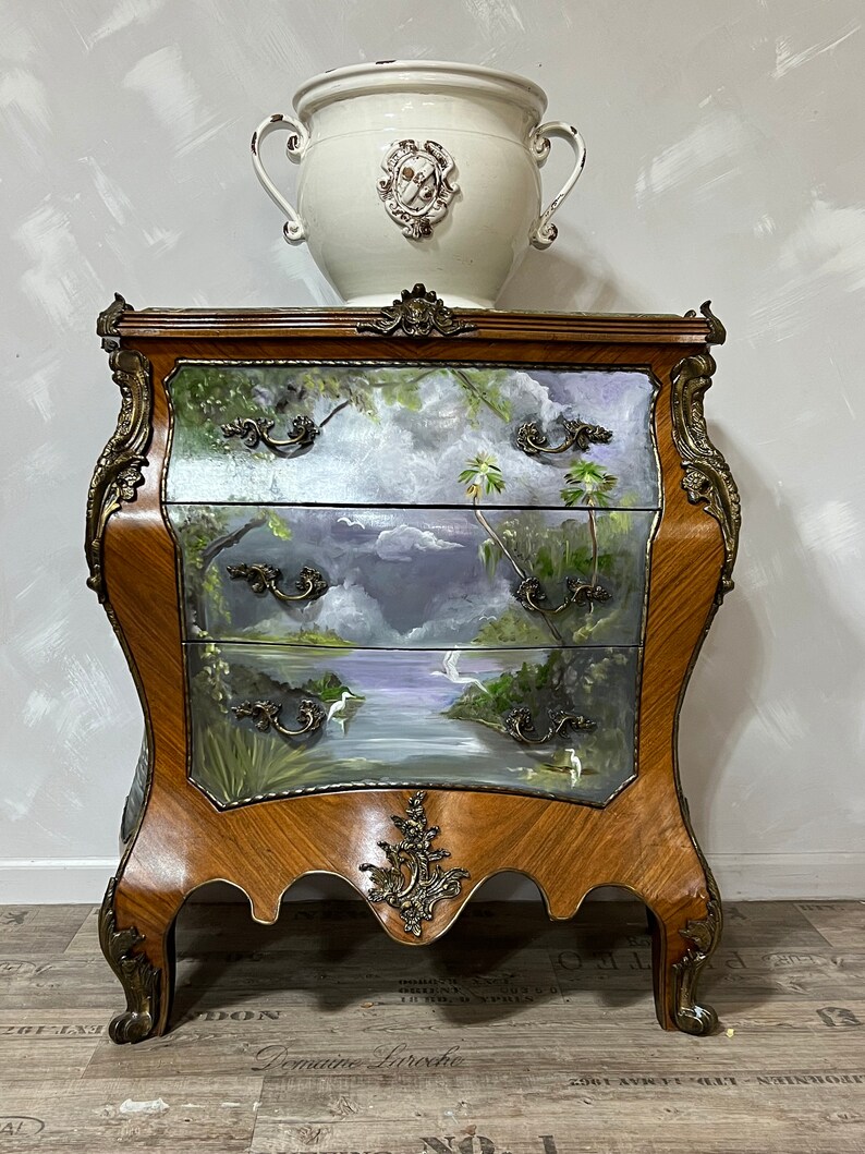 Pode incluir: Um ba&uacute; de gavetas de madeira com uma cena pintada de um lago e palmeiras. O ba&uacute; tem detalhes dourados ornamentados e est&aacute; sobre um piso de madeira. Um vaso de cer&acirc;mica branco com um design decorativo est&aacute; no topo do ba&uacute;.
