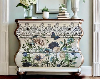 May include: A white, three-drawer dresser with a floral design and gold accents. The dresser has a curved, ornate top and sits on four legs.