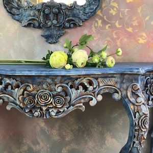 May include: A blue, ornate console table with carved details and a distressed finish. The table has a shelf and a decorative top with a floral design. The table is decorated with a bouquet of white and yellow flowers.