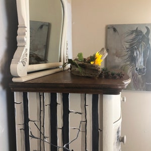 Puede incluir: Una c&oacute;moda blanca con un dise&ntilde;o de &aacute;rbol de abedul en blanco y negro. La c&oacute;moda tiene una tapa de madera y un espejo en la parte superior. Un peque&ntilde;o cuenco de madera con un girasol y vegetaci&oacute;n se encuentra en la c&oacute;moda.