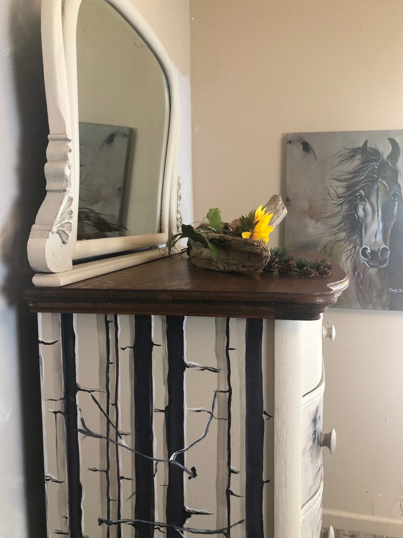 Puede incluir: Una c&oacute;moda blanca con un dise&ntilde;o de &aacute;rbol de abedul en blanco y negro. La c&oacute;moda tiene una tapa de madera y un espejo en la parte superior. Un peque&ntilde;o cuenco de madera con un girasol y vegetaci&oacute;n se encuentra en la c&oacute;moda.