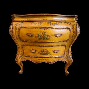 May include: A yellow antique chest of drawers with two drawers and ornate floral detailing. The chest has a curved front and legs, with bronze-colored trim and drawer pulls. The floral design includes red, orange, and green accents.