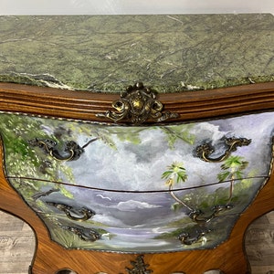 Pode incluir: Uma c&oacute;moda de tr&ecirc;s gavetas com tampo de m&aacute;rmore verde e uma cena de paisagem pintada &agrave; m&atilde;o na frente. A c&oacute;moda tem ferragens de lat&atilde;o ornamentadas e pernas de madeira esculpidas.