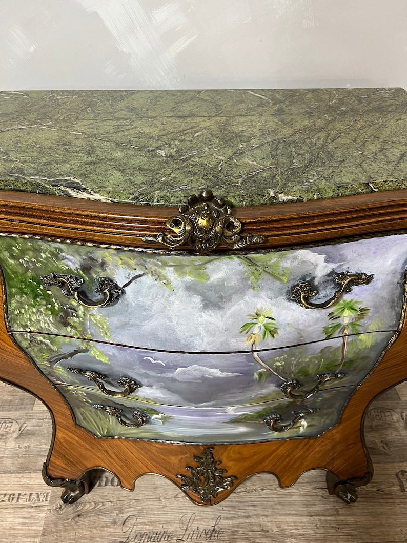 Pode incluir: Uma c&oacute;moda de tr&ecirc;s gavetas com tampo de m&aacute;rmore verde e uma cena de paisagem pintada &agrave; m&atilde;o na frente. A c&oacute;moda tem ferragens de lat&atilde;o ornamentadas e pernas de madeira esculpidas.