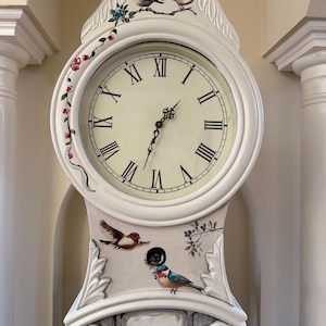 May include: A white grandfather clock with a cream-colored clock face and black Roman numerals. The clock is decorated with painted birds and floral accents in red, blue, and green. The clock hands point to approximately 7:00.