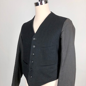 Puede incluir: Un chaleco de lana negro con un dobladillo puntiagudo y mangas de tela de algodón gris a contraste. El chaleco tiene un cuello en V y seis botones negros en la parte delantera.